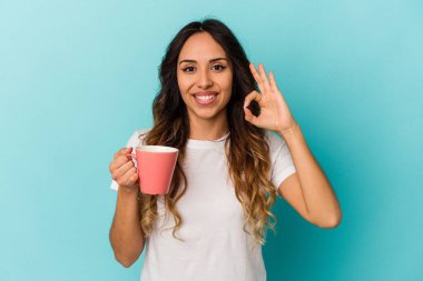 Elinde kupa tutan genç Meksikalı kadın mavi arka planda izole edilmiş neşeli ve kendinden emin..