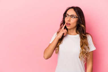 Pembe arka planda izole edilmiş genç Meksikalı kadın şüpheli ve şüpheci bir ifadeyle yana bakıyor..