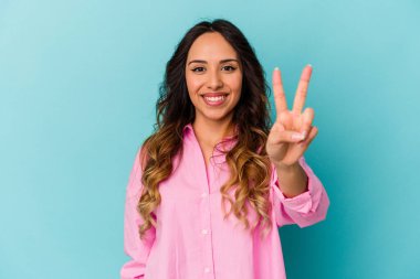 Mavi arka planda izole edilmiş genç Meksikalı kadın zafer işareti gösteriyor ve gülümsüyor..