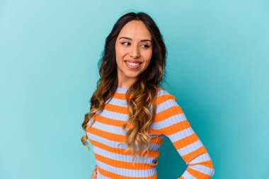 Mavi arka planda izole edilmiş genç Meksikalı kadın mutlu bir şekilde gülüyor ve ellerini karnının üstünde tutarak eğleniyor..