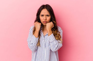 Pembe arka planda izole edilmiş genç Meksikalı kadın yumruğunu kameraya gösteriyor, agresif yüz ifadesi..