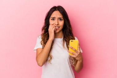 Elinde cep telefonu olan genç Meksikalı kadın pembe arka planda izole edilmiş tırnak yiyor, gergin ve endişeli..