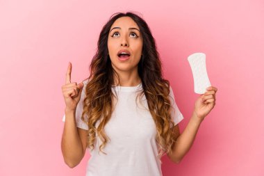 Genç Meksikalı bir kadın pembe arka plana izole edilmiş bir kompres tutuyor ve ağzı açık bir şekilde üst tarafı işaret ediyor..