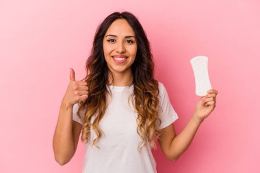 Pembe arka planda bir kompres tutan genç Meksikalı kadın gülümsüyor ve başparmağını kaldırıyor.