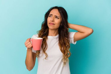 Genç Meksikalı bir kadın mavi arka planda bir kupa tutuyor. Kafasının arkasına dokunuyor, düşünüyor ve bir seçim yapıyor..