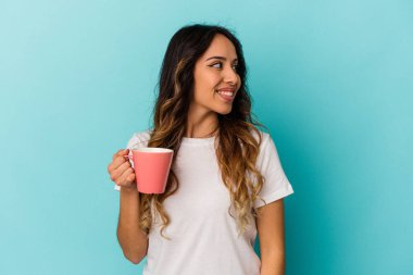 Mavi arka planda bir kupa tutan genç Meksikalı kadın gülümsüyor, neşeli ve keyifli görünüyor..