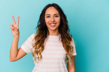Mavi arka planda izole edilmiş genç Meksikalı kadın zafer işareti gösteriyor ve gülümsüyor..