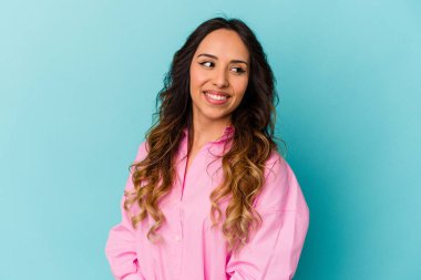 Mavi arka planda izole edilmiş genç Meksikalı kadın gülümseyen, neşeli ve keyifli bir kenara bakıyor..
