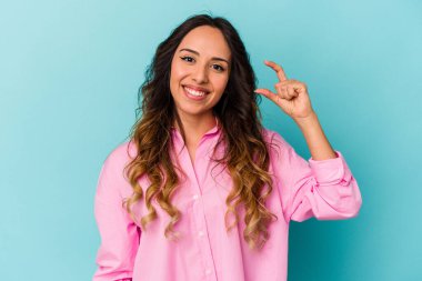 Young mexican woman isolated on blue background holding something little with forefingers, smiling and confident. clipart