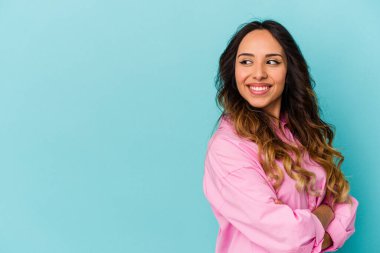 Mavi arka planda izole edilmiş genç Meksikalı kadın kollarını kavuşturarak gülümsüyor..