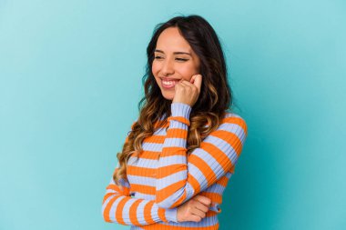 Mavi arka planda izole edilmiş genç Meksikalı kadın mutlu ve kendinden emin gülümsüyor, elleriyle çenesine dokunuyor..