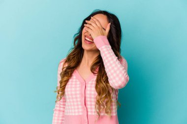 Mavi arka planda izole edilmiş genç Meksikalı kadın elleriyle gözlerini kapatıyor, gülümseyerek bir sürpriz bekliyor..