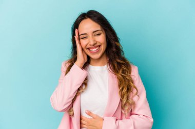 Mavi arka planda izole edilmiş genç Meksikalı kadın mutlu bir şekilde gülüyor ve ellerini karnının üstünde tutarak eğleniyor..