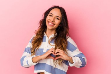 Pembe arka planda izole edilmiş genç Meksikalı kadın gülümsüyor ve elleriyle kalp şeklini gösteriyor..