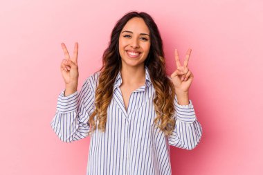 Pembe arka planda izole edilmiş genç Meksikalı kadın zafer işareti gösteriyor ve gülümsüyor..