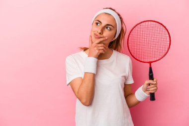 Pembe arka planda izole edilmiş badminton raketi tutan beyaz bir kadın şüpheli ve şüpheci bir ifadeyle yana bakıyor..