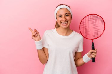 Genç, sarışın, beyaz bir kadın elinde bir badminton raketi tutuyor pembe arka planda izole edilmiş gülümsüyor ve boş alanda bir şeyler gösteriyor..