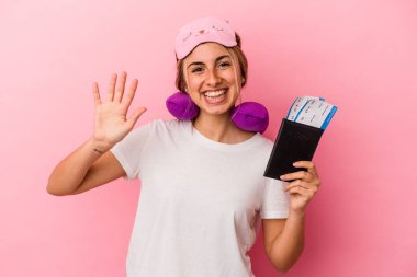 Elinde pasaport ve seyahat biletleri olan beyaz saçlı genç bir kadın pembe arka planda tek başına gülümseyerek beş numarayı parmaklarıyla gösteriyor..