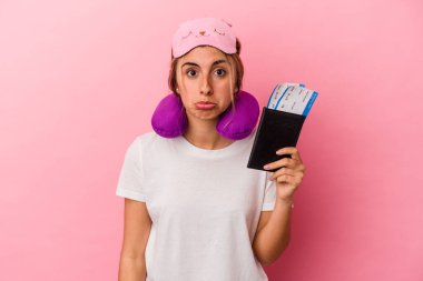 Pasaport ve seyahat biletleri taşıyan beyaz, genç bir kadın pembe arka planda tek başına omuz silkiyor ve gözleri açık..