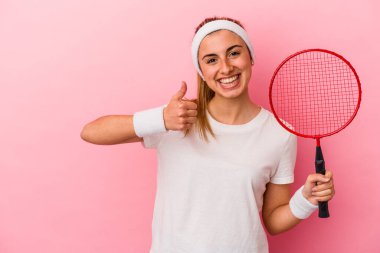 Genç, sarışın, beyaz bir kadın elinde badminton raketi tutuyor pembe arka planda izole edilmiş gülümsüyor ve başparmağını kaldırıyor.