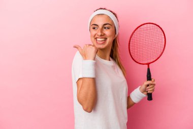 Genç, sarışın, beyaz bir kadın elinde badminton raketi tutuyor. Pembe arka planda soyutlanmış, baş parmağı uzakta, güler yüzlü ve kaygısız..