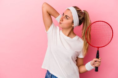 Genç, sarışın, beyaz bir kadın elinde bir badminton raketi tutuyor pembe arka planda izole edilmiş kafasının arkasına dokunuyor, düşünüyor ve bir seçim yapıyor..