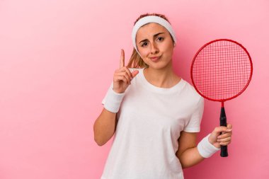 Genç, sarışın, beyaz bir kadın elinde badminton raketi tutuyor. Pembe arka planda izole edilmiş. Parmağıyla bir numarayı gösteriyor..