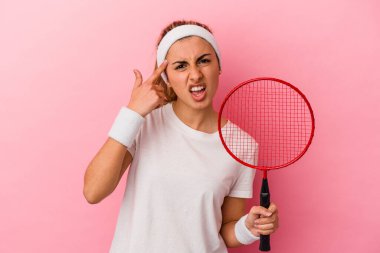 Genç, sarışın, beyaz bir kadın elinde badminton raketi tutuyor. Pembe arka planda izole edilmiş. İşaret parmağıyla yaptığı hayal kırıklığını gösteriyor..