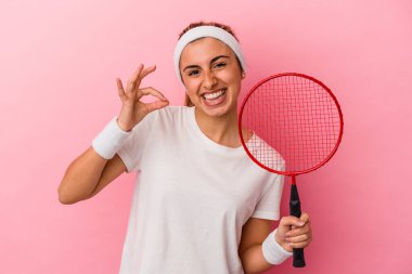 Genç, sarışın, beyaz bir kadın elinde badminton raketi tutuyor. Pembe arka planda izole edilmiş. Neşeli ve kendinden emin..