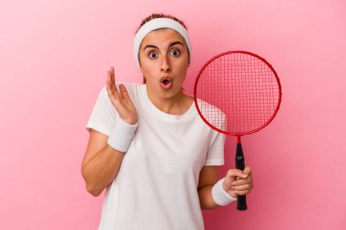 Genç, sarışın, beyaz bir kadın elinde badminton raketi tutuyor. Pembe arka planda izole edilmiş. Şaşırmış ve şok olmuş..