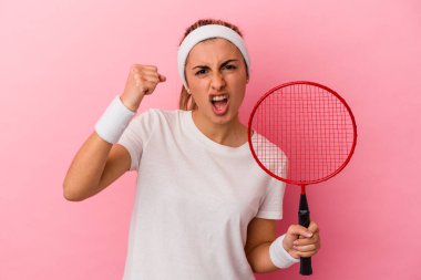 Genç, sarışın, beyaz bir kadın elinde badminton raketi tutuyor. Pembe arka planda izole edilmiş. Kameraya yumruğunu gösteriyor..