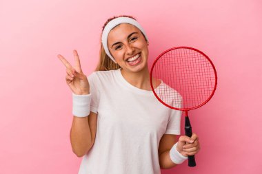 Genç, sarışın, beyaz bir kadın elinde bir badminton raketi tutuyor pembe arka planda izole edilmiş neşeli ve kaygısız parmaklarıyla barış sembolü gösteriyor..