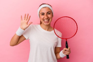 Genç, sarışın, beyaz bir kadın elinde badminton raketi tutuyor pembe arka planda izole edilmiş, gülümseyen beş numarayı parmaklarıyla gösteriyor..