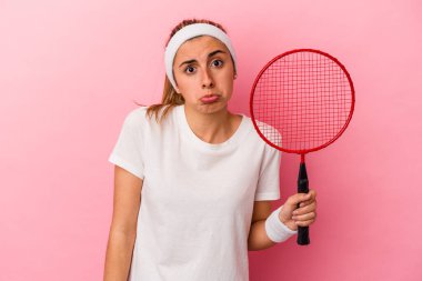 Pembe arka planda izole edilmiş badminton raketi tutan genç sarışın beyaz kadın omuzlarını silkeliyor ve gözleri açık..
