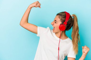 Kulaklıkla müzik dinleyen genç sarışın kadın mavi arka planda izole edilmiş zafer ve kazanma kavramından sonra yumruğunu kaldırıyor..