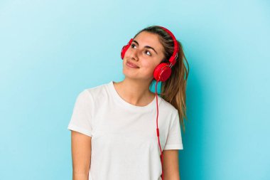 Genç sarışın kadın kulaklıkla müzik dinliyor mavi arka planda izole edilmiş hedeflere ve amaçlara ulaşma hayali kuruyor.