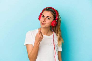 Genç sarışın kadın kulaklıkla müzik dinliyor. Mavi arka planda izole edilmiş. Seni işaret ediyor..