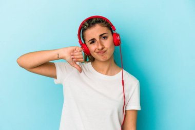 Kulaklıkla müzik dinleyen genç sarışın bir kadın. Mavi arka planda izole edilmiş. Sevimsiz bir jest gösteriyor. Anlaşmazlık konsepti.