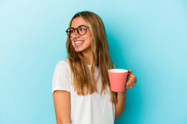 Mavi arka planda saklanmış bir kupa tutan beyaz saçlı genç bir kadın gülümsüyor, neşeli ve keyifli görünüyor..