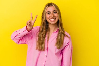 Sarı arka planda izole edilmiş genç sarışın kadın zafer işareti gösteriyor ve gülümsüyor..