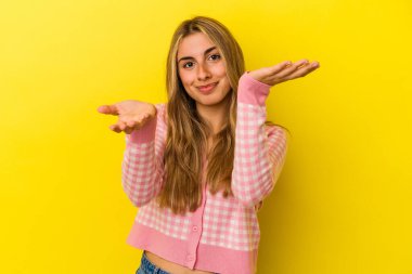 Sarı arka planda izole edilmiş genç sarışın kadın kollarıyla ölçeği yapıyor, mutlu ve kendinden emin hissediyor..