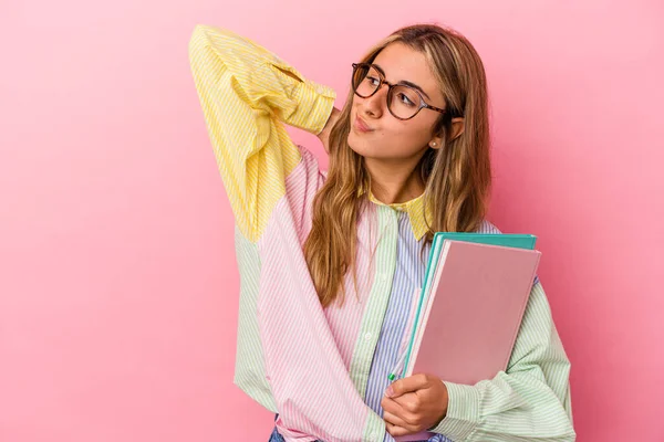 Young caucasian blonde student woman holding books isolated touching back of head, thinking and making a choice.