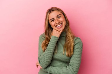 Genç sarışın beyaz kadın pembe arka planda izole edilmiş mutlu ve kendinden emin gülümsüyor, elleriyle çenesine dokunuyor..