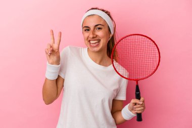 Genç, sarışın, beyaz bir kadın elinde badminton raketi tutuyor. Pembe arka planda iki numaranın parmakları olduğunu gösteriyor..