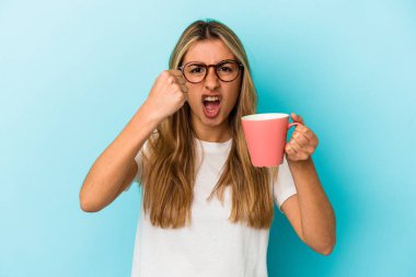 Mavi arka planda izole edilmiş bir kupa tutan beyaz, genç bir kadın kameraya yumruğunu gösteriyor, agresif bir yüz ifadesi..