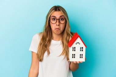 Young caucasian woman holding a house model isolated on blue background shrugs shoulders and open eyes confused. clipart