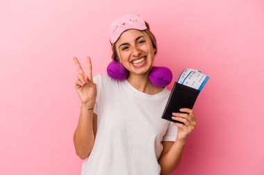 Elinde pasaport ve seyahat biletleri olan beyaz saçlı genç bir kadın pembe arka planda izole edilmiş neşeli ve tasasız bir barış sembolü gösteriyor..