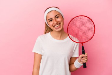 Genç, sarışın, beyaz bir kadın elinde badminton raketi tutuyor pembe arka planda izole edilmiş mutlu, güler yüzlü ve neşeli..
