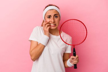Genç, sarışın, beyaz bir kadın elinde badminton raketi tutuyor. Pembe arka planda izole edilmiş, tırnaklarını yiyor. Gergin ve endişeli..