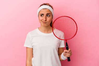 Pembe arka planda izole edilmiş badminton raketi tutan genç, sarışın, beyaz bir kadın. Kafası karışmış ve kendinden emin değil..